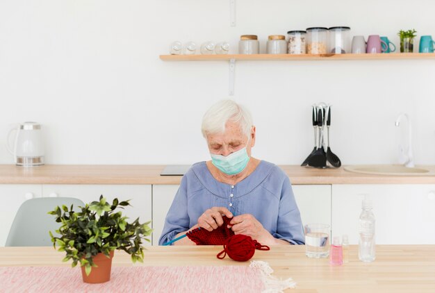 Vista frontale della donna più anziana con la maschera medica che tricotta a casa