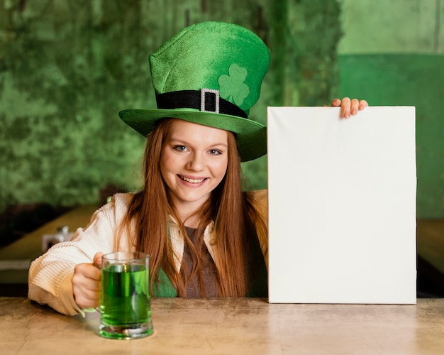 Vista frontale della donna sorridente che celebra st. patrick's day al bar con cartello bianco e drink