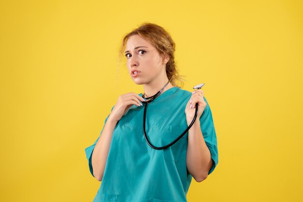 Vista frontale della dottoressa in tuta medica con lo stetoscopio sulla parete gialla