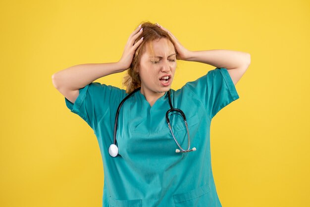 Vista frontale della dottoressa in tuta medica con lo stetoscopio sulla parete gialla