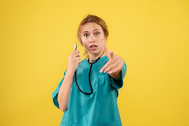 Vista frontale della dottoressa in tuta medica con lo stetoscopio sulla parete gialla