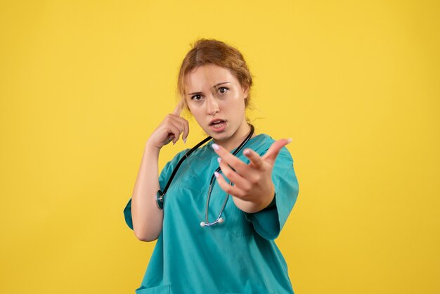 Vista frontale della dottoressa in tuta medica con lo stetoscopio sulla parete gialla