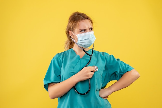 Vista frontale della dottoressa in tuta medica e maschera con lo stetoscopio sulla parete gialla
