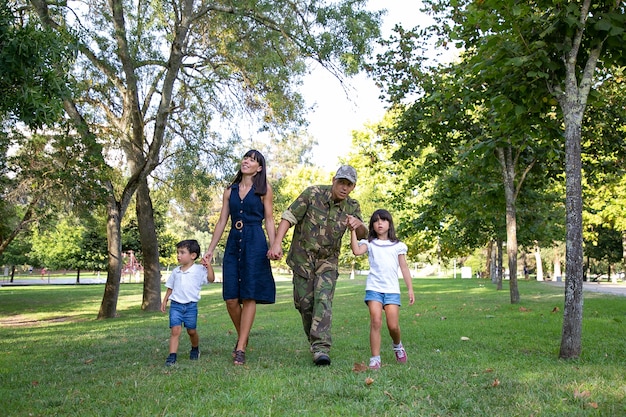 Vista frontale della famiglia felice che cammina insieme sul prato nel parco. Padre che indossa l'uniforme militare e mostra qualcosa alla figlia. Mamma dai capelli lunghi che sorride. Ricongiungimento familiare e concetto di ritorno a casa