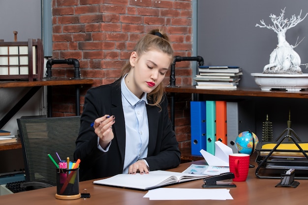 Vista frontale della giovane assistente femminile sicura di sé seduta alla sua scrivania e leggendo un documento in ufficio