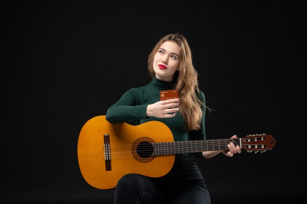 Vista frontale della giovane bella musicista sognante che tiene in mano chitarra e carta di credito al buio