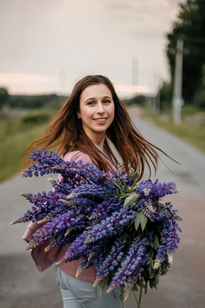 Vista frontale della giovane bella ragazza bruna in piedi sulla strada suburbana con un grande mazzo di lupini viola selvatici