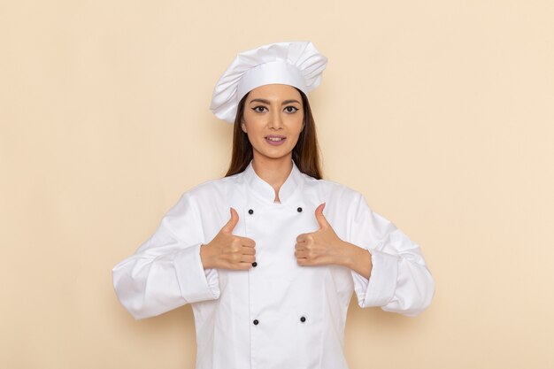 Vista frontale della giovane cuoca in tuta bianca da cuoco che sorride e mostra segni simili sul muro bianco chiaro