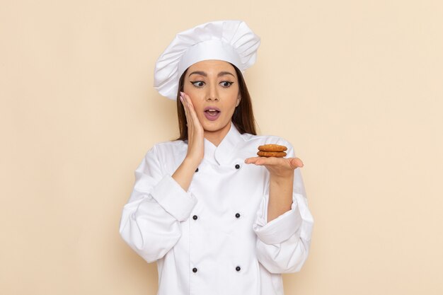 Vista frontale della giovane cuoca in vestito bianco da cuoco che tiene piccoli biscotti sulla parete bianco-chiaro