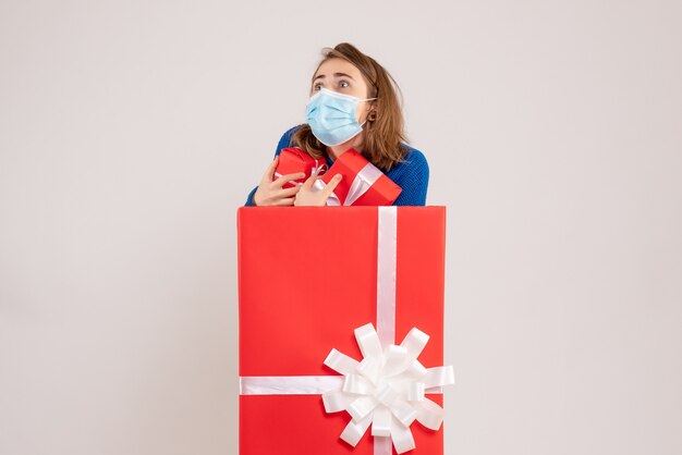 Vista frontale della giovane donna all'interno della confezione regalo in maschera con regali sul muro bianco
