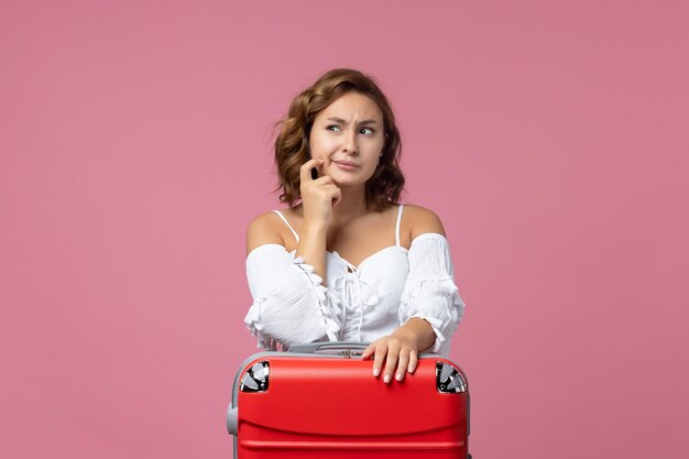 Vista frontale della giovane donna che pensa e si prepara per il viaggio con la borsa rossa sul muro rosa