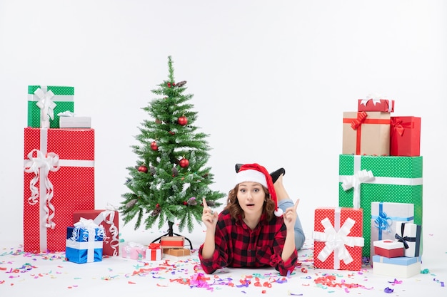 Vista frontale della giovane donna che posa intorno ai regali di Natale e al piccolo albero di festa sulla parete bianca