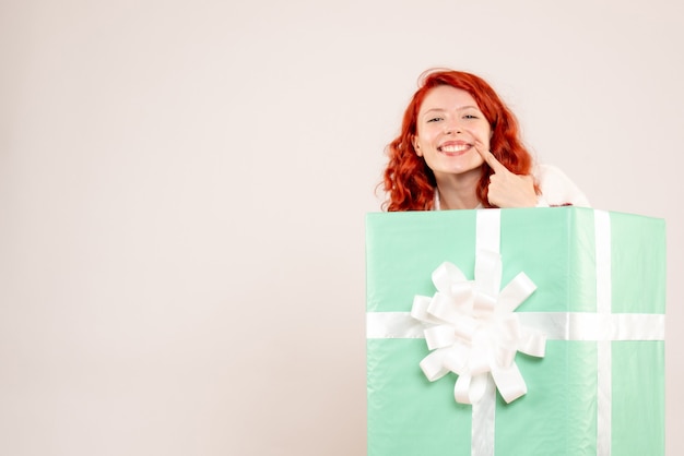 Vista frontale della giovane donna che si nasconde dentro il presente sorridente sul muro bianco