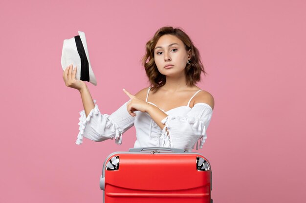 Vista frontale della giovane donna che si prepara per le vacanze e tiene il cappello sul muro rosa