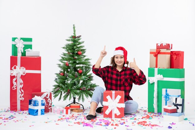 Vista frontale della giovane donna che si siede intorno ai regali e al piccolo albero di festa sulla parete bianca