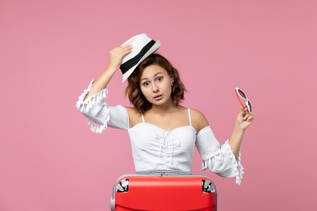 Vista frontale della giovane donna che tiene in mano cappello e occhiali da sole con borsa rossa su un viaggio di viaggio di vacanza modello di mare di colore rosa