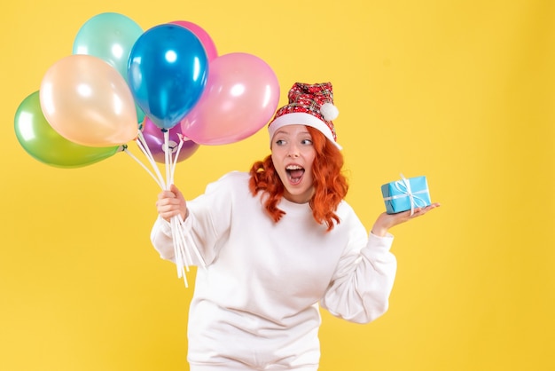 Vista frontale della giovane donna che tiene poco presente e palloncini colorati sulla parete gialla