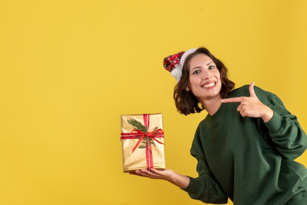 Vista frontale della giovane donna che tiene poco regalo di Natale sulla parete gialla