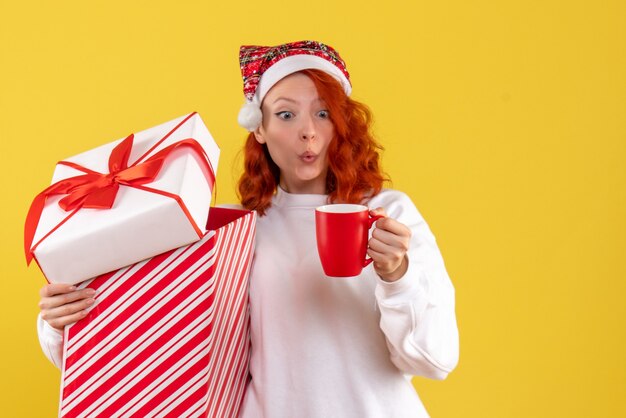 Vista frontale della giovane donna che tiene regalo di Natale e tazza di tè sulla parete gialla