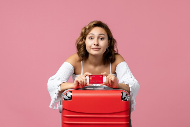 Vista frontale della giovane donna con la borsa delle vacanze che tiene la carta di credito sulla parete rosa