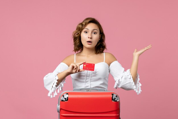 Vista frontale della giovane donna con la borsa delle vacanze che tiene la carta di credito sulla parete rosa