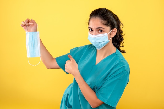 Vista frontale della giovane donna con maschera medica che tiene la maschera che fa il pollice sul segno sulla parete gialla