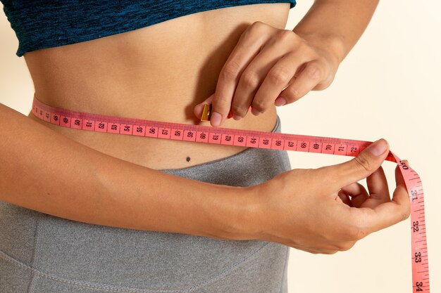 Vista frontale della giovane donna con un corpo in forma in camicia che misura la sua vita sul muro bianco