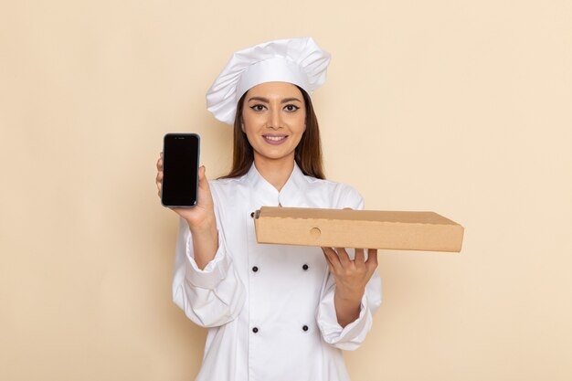 Vista frontale della giovane donna cuoca in vestito bianco da cuoco che tiene il telefono e la scatola di cibo sulla parete bianco-chiaro