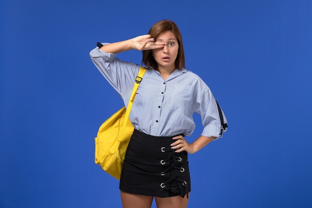 Vista frontale della giovane donna in camicia blu gonna nera che indossa uno zaino giallo in posa sulla parete blu chiaro