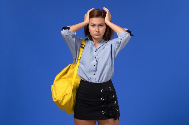 Vista frontale della giovane donna in camicia blu gonna nera che indossa uno zaino giallo in posa sulla parete blu chiaro