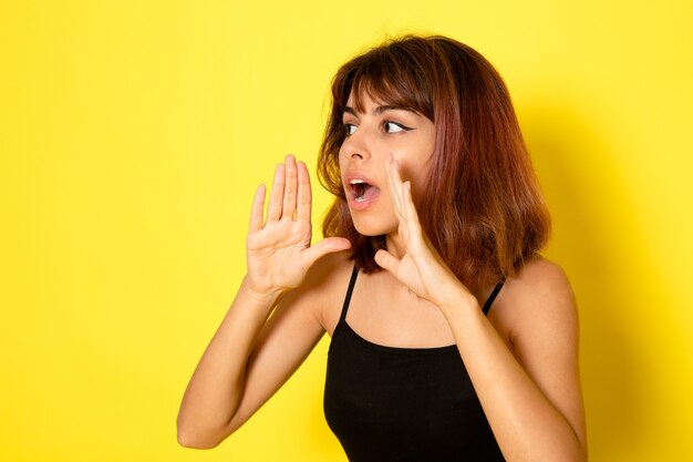 Vista frontale della giovane donna in camicia nera che bisbiglia e chiama sul muro giallo chiaro