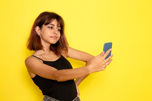 Vista frontale della giovane donna in camicia nera che cattura un selfie sulla parete giallo chiaro