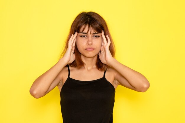 Vista frontale della giovane donna in camicia nera e jeans grigi che hanno mal di testa sulla parete gialla