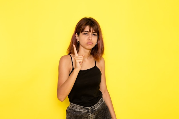 Vista frontale della giovane donna in camicia nera e jeans grigi che minacciano con il dito sulla parete gialla