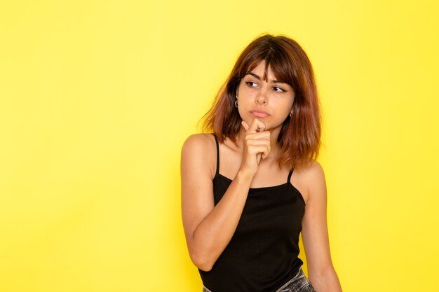 Vista frontale della giovane donna in camicia nera e jeans grigi pensando sulla parete gialla