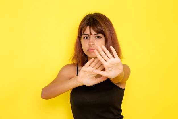 Vista frontale della giovane donna in camicia nera e jeans grigi prudenti sulla parete gialla