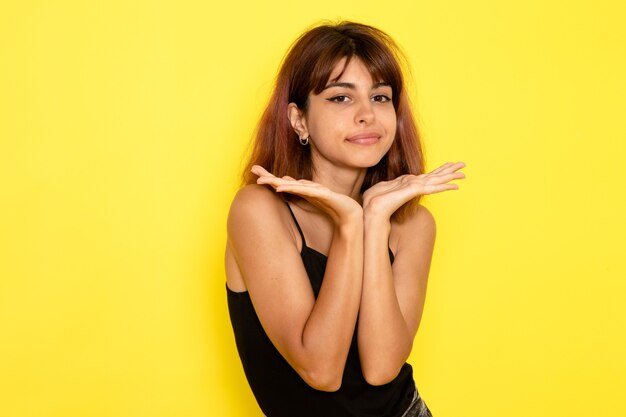 Vista frontale della giovane donna in camicia nera in posa con espressione carina sulla parete giallo chiaro