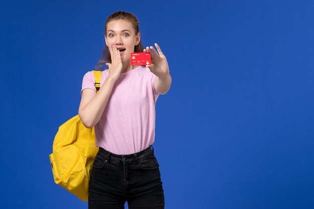 Vista frontale della giovane donna in maglietta rosa che indossa lo zaino giallo che tiene il cartellino rosso di plastica sulla parete blu