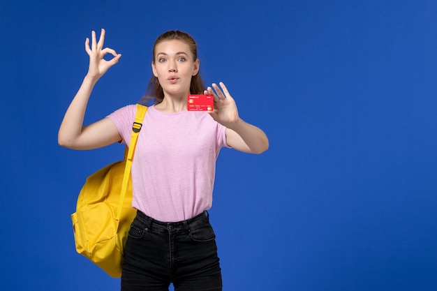 Vista frontale della giovane donna in maglietta rosa che indossa lo zaino giallo che tiene il cartellino rosso di plastica sulla parete blu