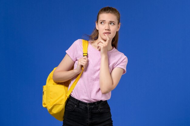 Vista frontale della giovane donna in maglietta rosa che indossa lo zaino giallo pensando sulla parete azzurra