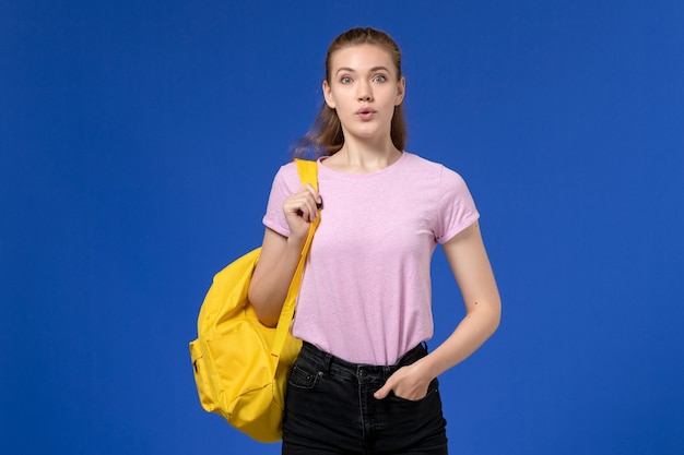 Vista frontale della giovane donna in maglietta rosa che indossa uno zaino giallo in piedi sulla parete blu