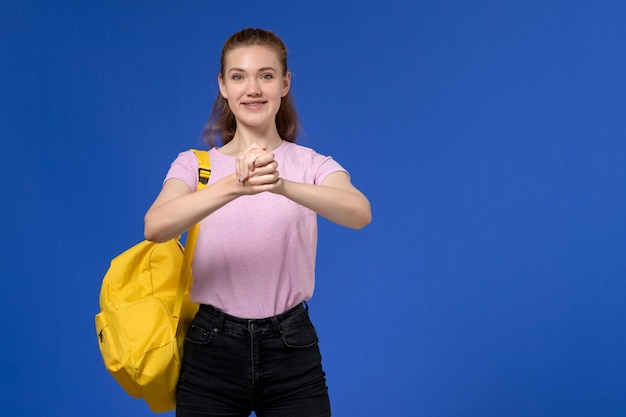 Vista frontale della giovane donna in maglietta rosa che indossa uno zaino giallo sulla parete blu