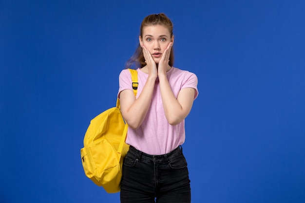 Vista frontale della giovane donna in maglietta rosa che indossa zaino giallo in posa sulla parete blu