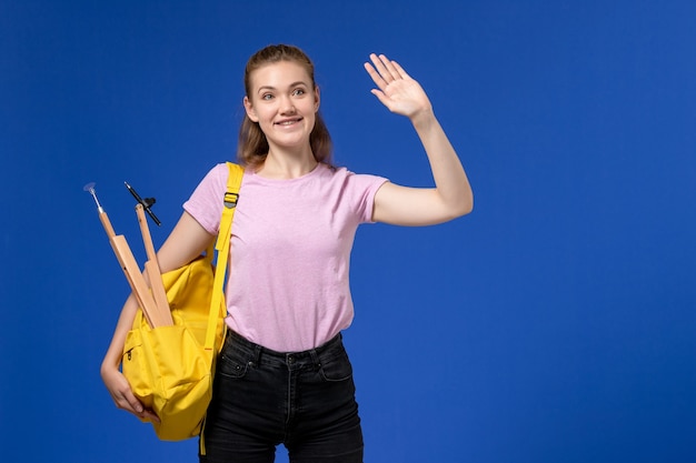 Vista frontale della giovane donna in maglietta rosa che indossa zaino giallo sorridente sulla parete blu