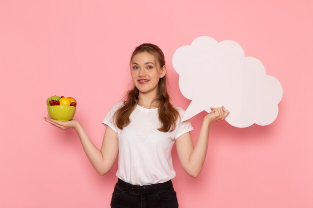 Vista frontale della giovane donna in t-shirt bianca che tiene piatto con frutta e segno bianco sulla parete rosa