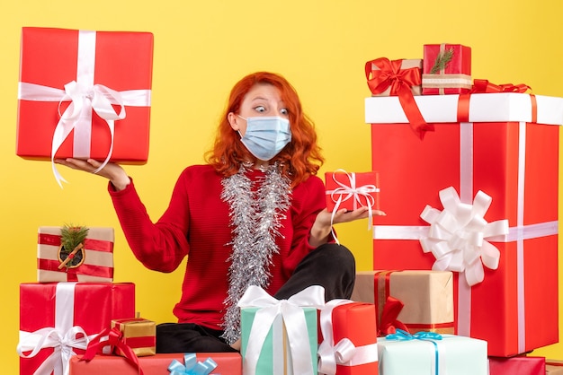 Vista frontale della giovane donna seduta intorno a regali di Natale in maschera su una parete gialla