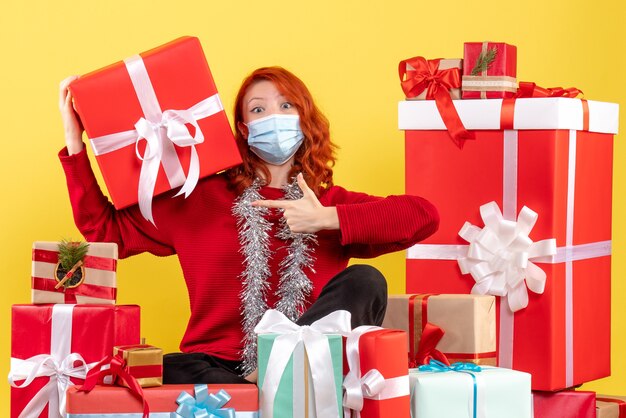 Vista frontale della giovane donna seduta intorno a regali di Natale in maschera su una parete gialla