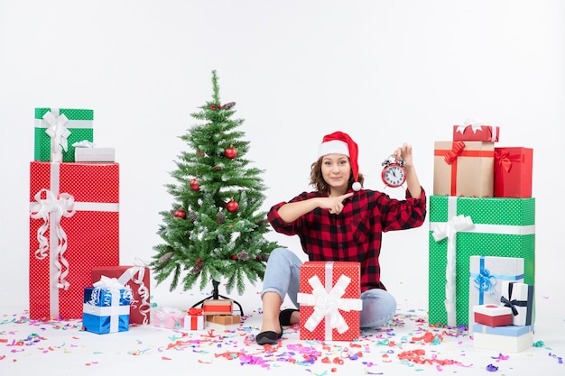 Vista frontale della giovane donna seduta intorno a regali di Natale in possesso di orologi sul muro bianco