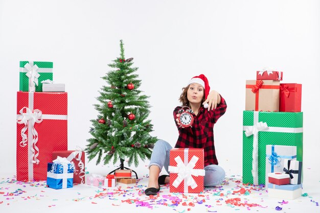 Vista frontale della giovane donna seduta intorno a regali di Natale in possesso di orologi sul muro bianco