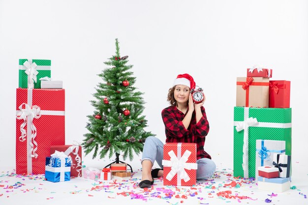 Vista frontale della giovane donna seduta intorno a regali di Natale in possesso di orologi sul muro bianco